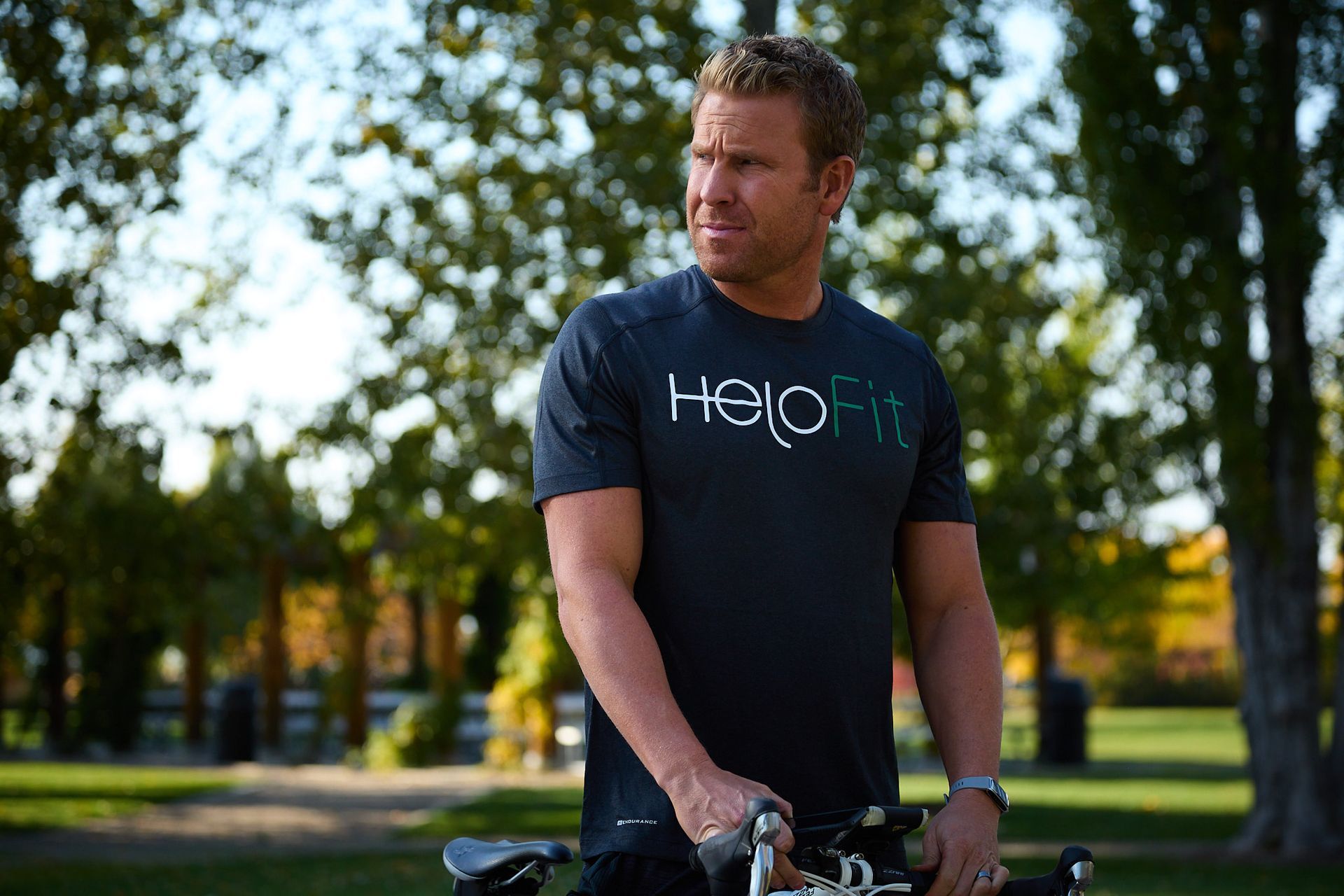 A person wearing a dark grey HeloFit t-shirt standing with a bicycle in a sunlit park.