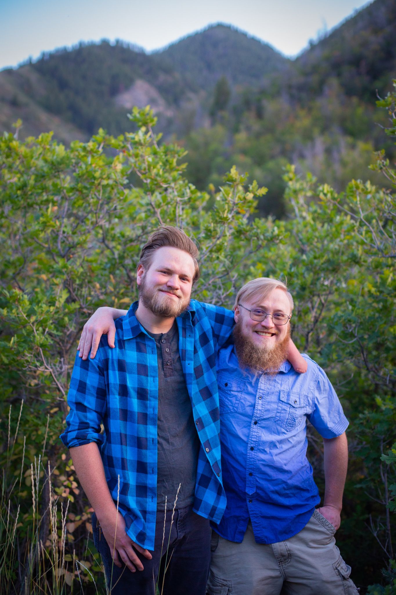 Two people with beards standing arm-in-arm in front of a green mountain landscape.