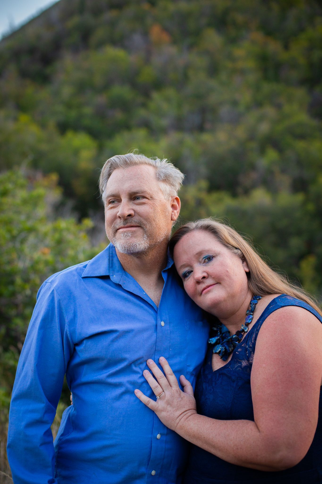 A man in a blue shirt and a woman in a navy dress pose together outdoors with a blurred, treed hill in the background.