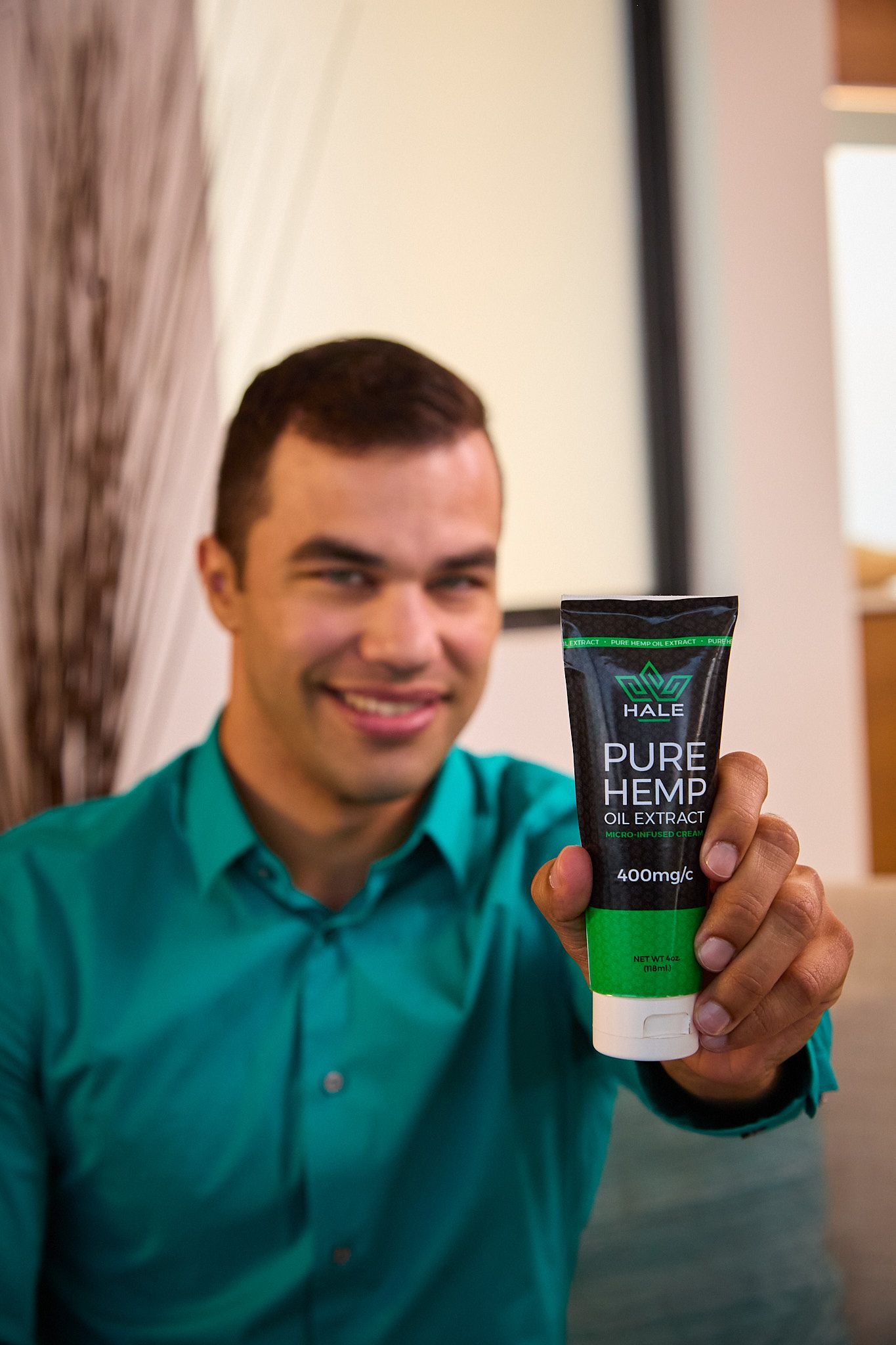 A smiling person in a teal shirt holding up a tube of Pure Hemp cream toward the camera.