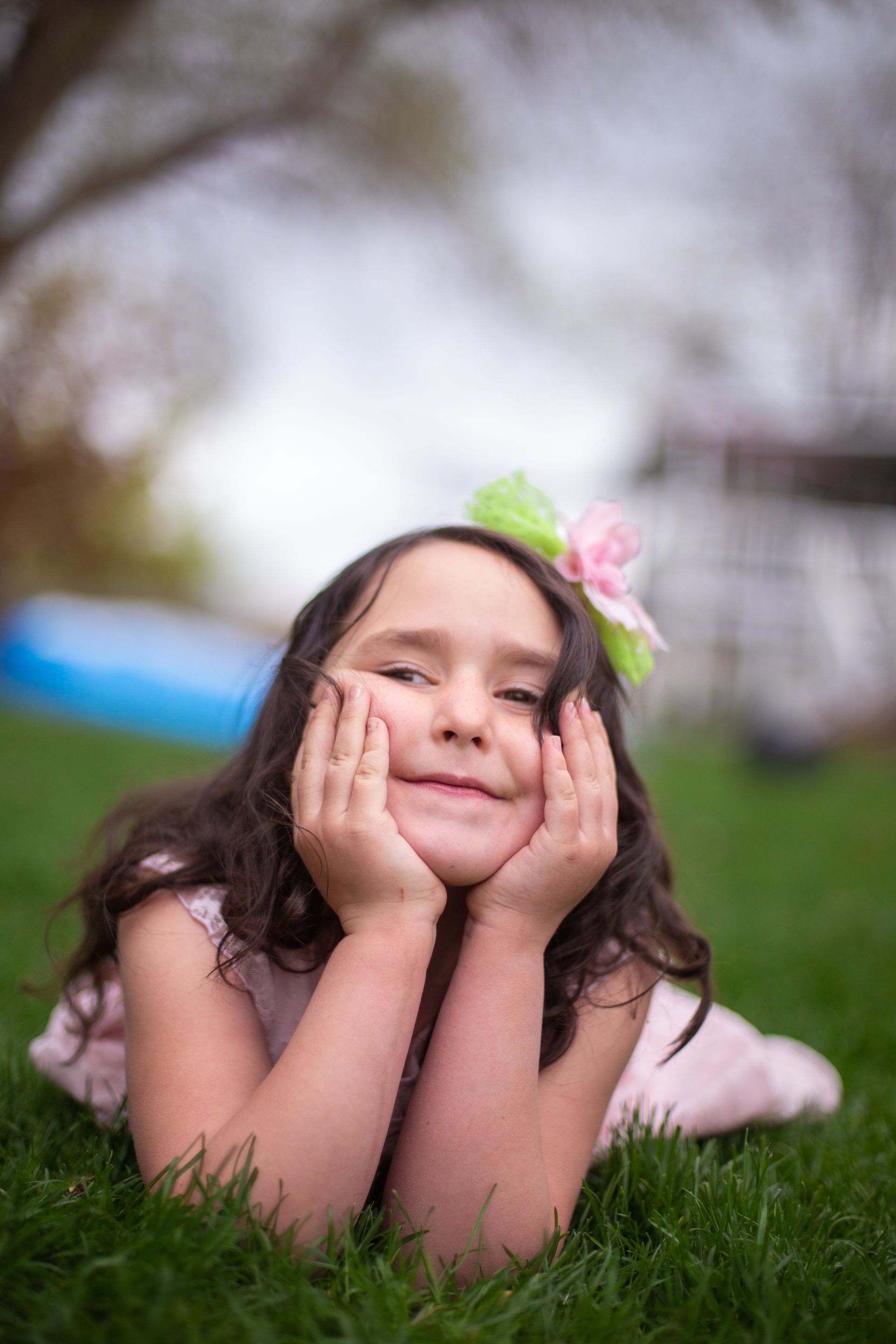 A person lying in the grass rests their chin on their hands, smiling, wearing a light pink dress and a floral hairpiece.