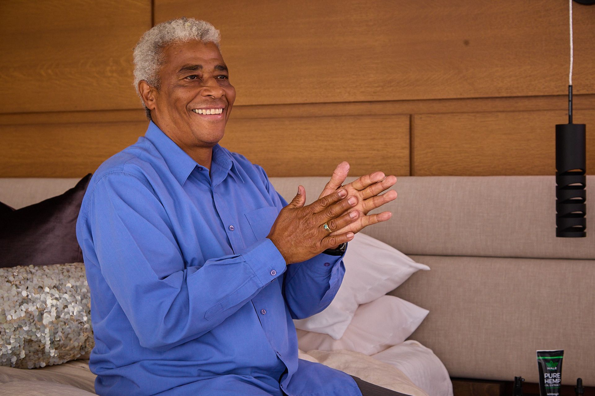 A person in a blue button-down shirt smiles and claps their hands while sitting on a bed in a modern, wood-paneled room.