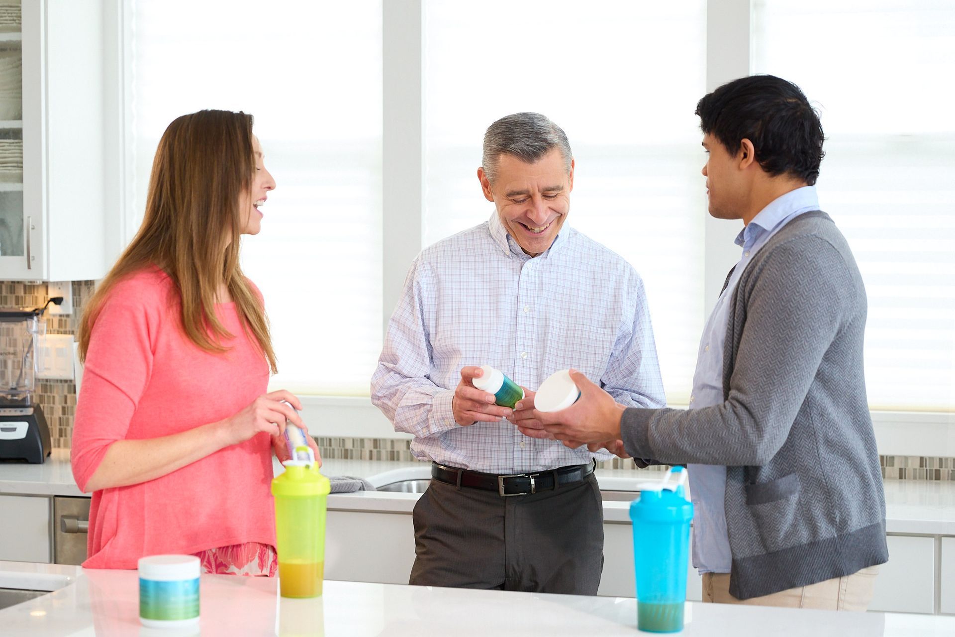 Three people in a kitchen stand around a counter, talking and holding supplements and smoothie cups.
