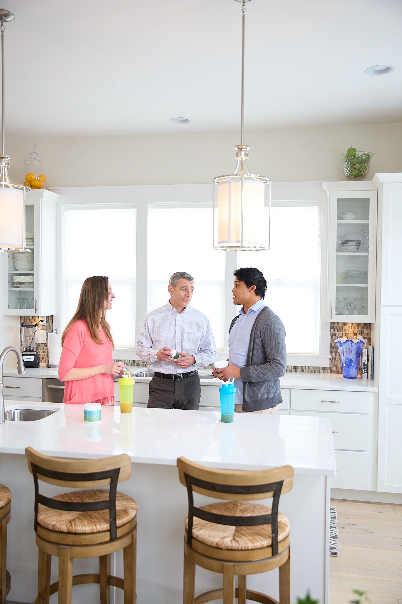 Three people stand in a bright, modern kitchen, talking and holding shaker bottles near a white island with bar stools.