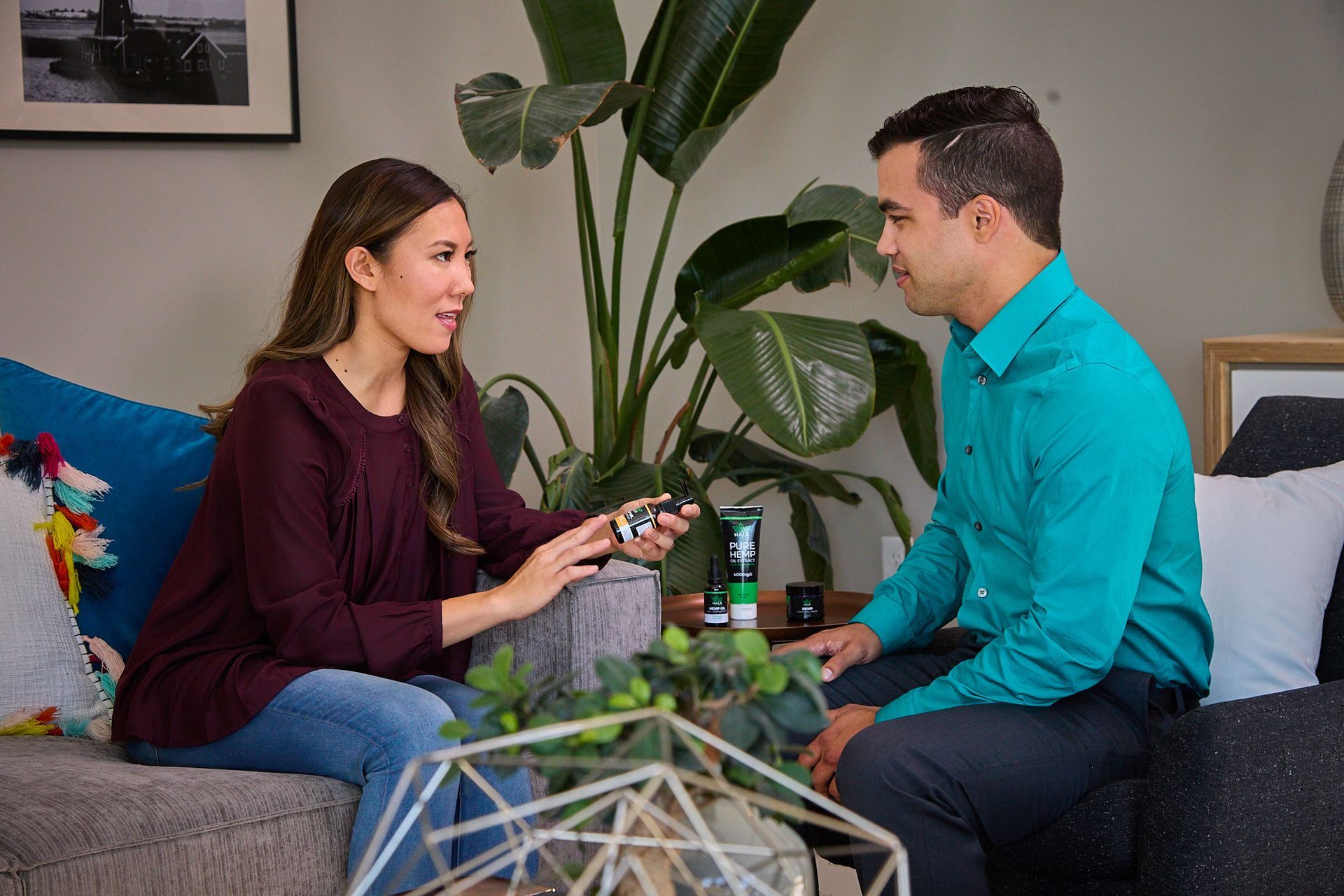 A person in a maroon shirt shows a product to another person in a teal shirt while sitting together on a couch.