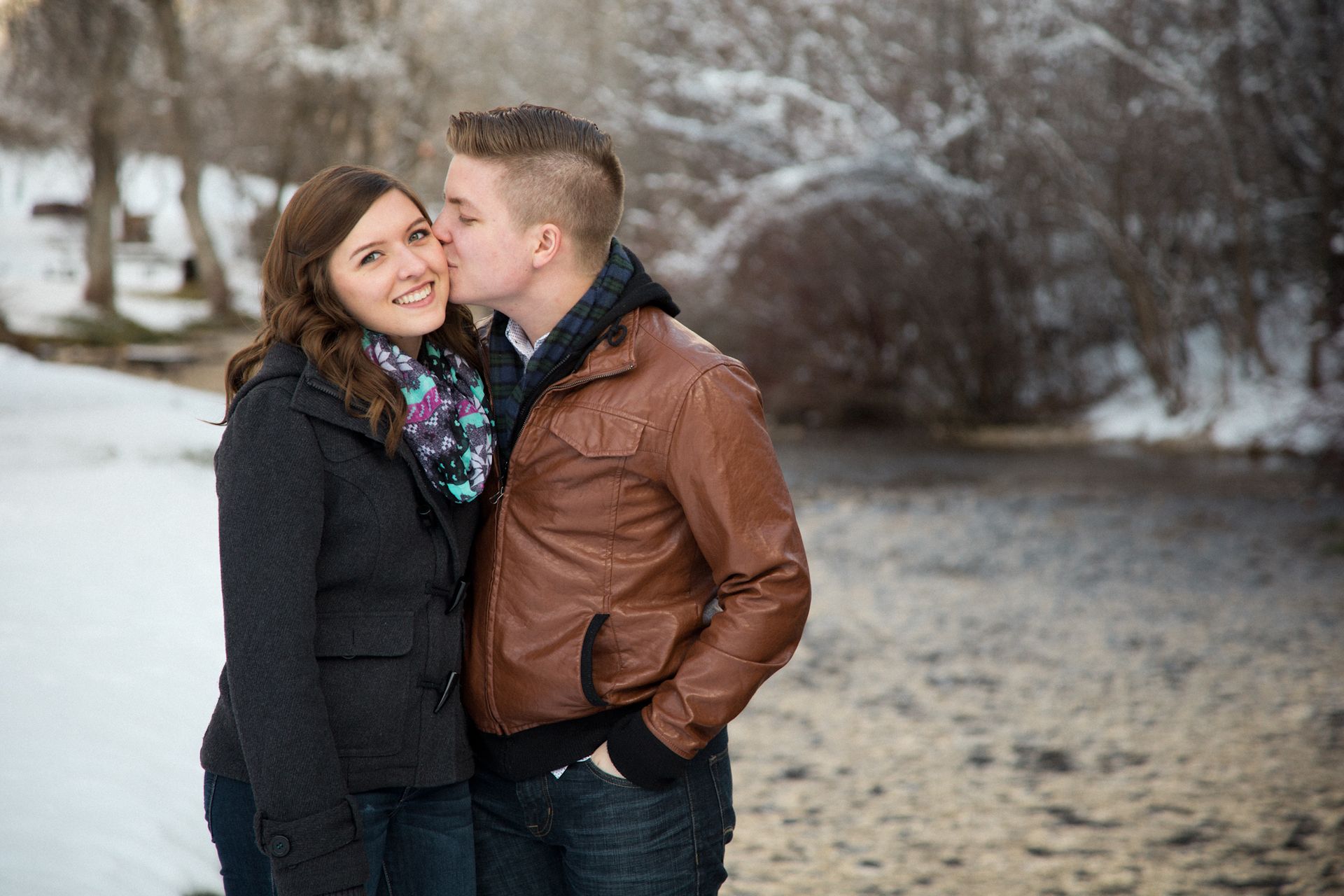 A person kisses their partner on the cheek outdoors in a snowy, wooded area beside a stream.