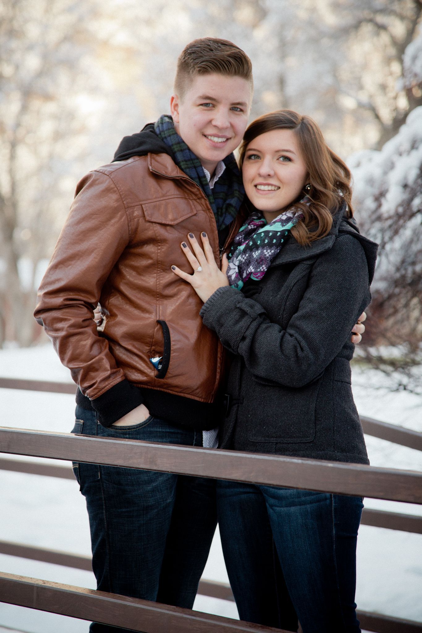A couple poses together on a wooden bridge in a snowy, outdoor setting, both wearing winter jackets.