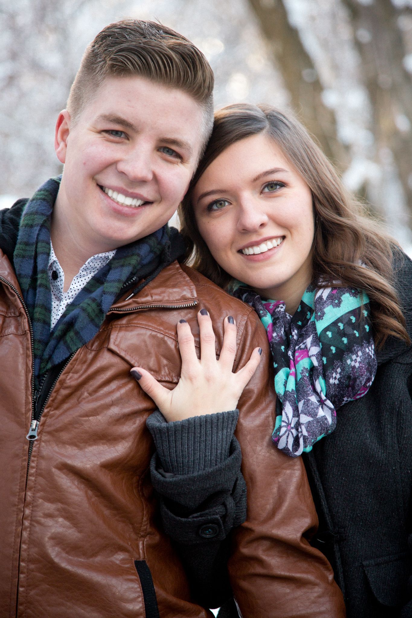 A smiling couple stands close outdoors in winter, wearing scarves and coats, with the woman's hand resting on the man's arm.