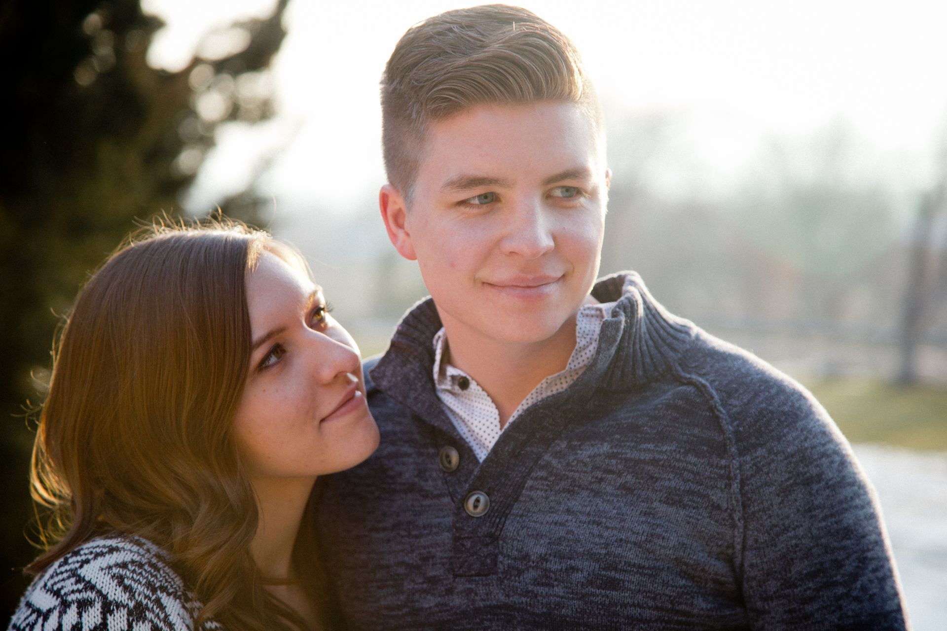 A person looks affectionately at a man standing beside them, both posing outdoors in soft, sunlit lighting.