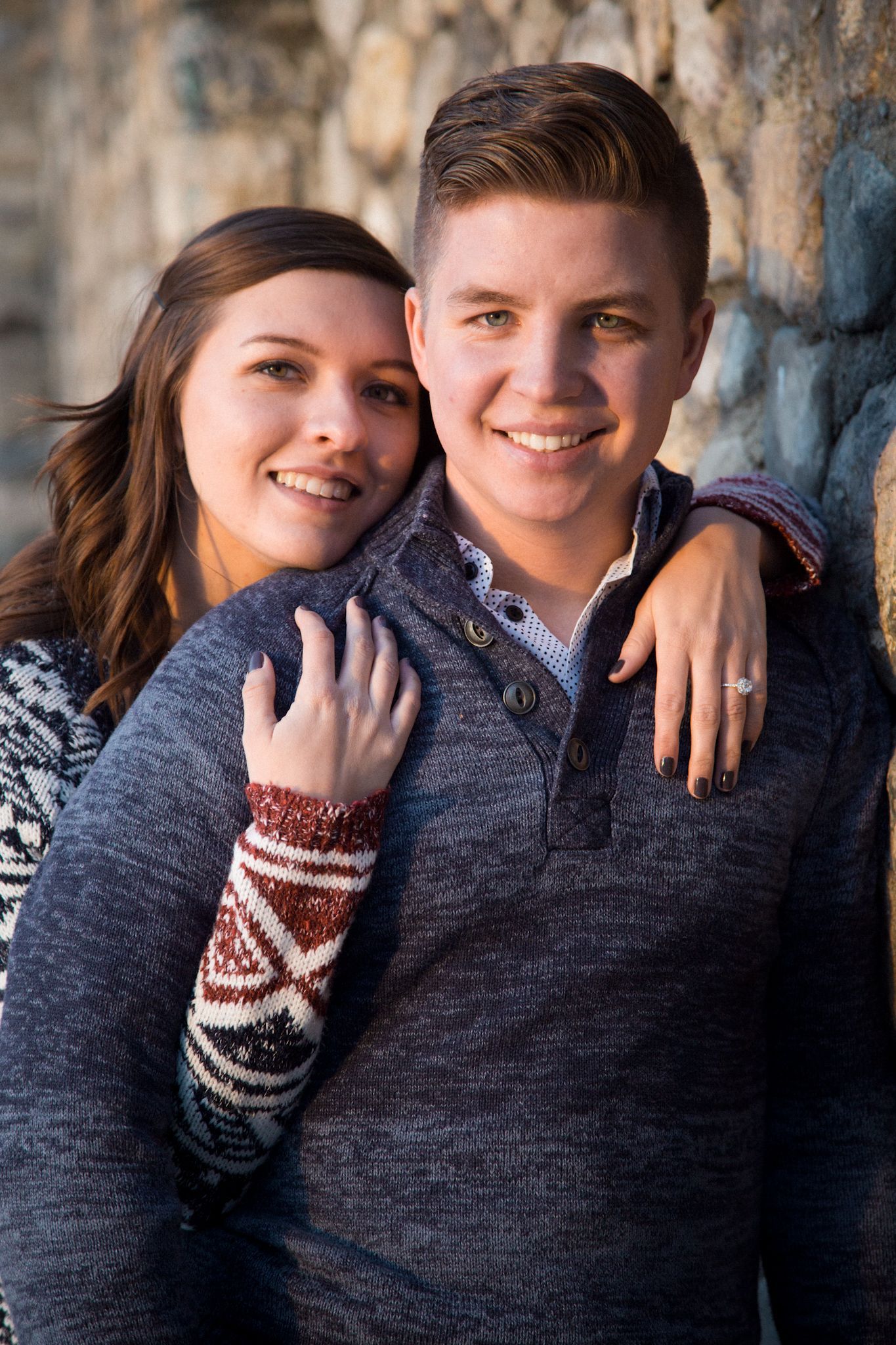 A smiling couple embraces against a stone wall; the woman rests her head on the man's shoulder.