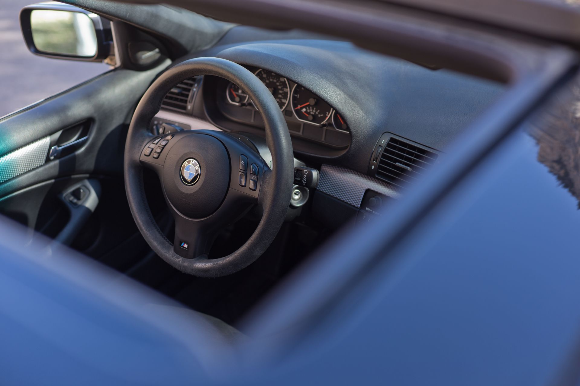 The driver's side interior of a BMW car, featuring a black steering wheel, dashboard, and side mirror.