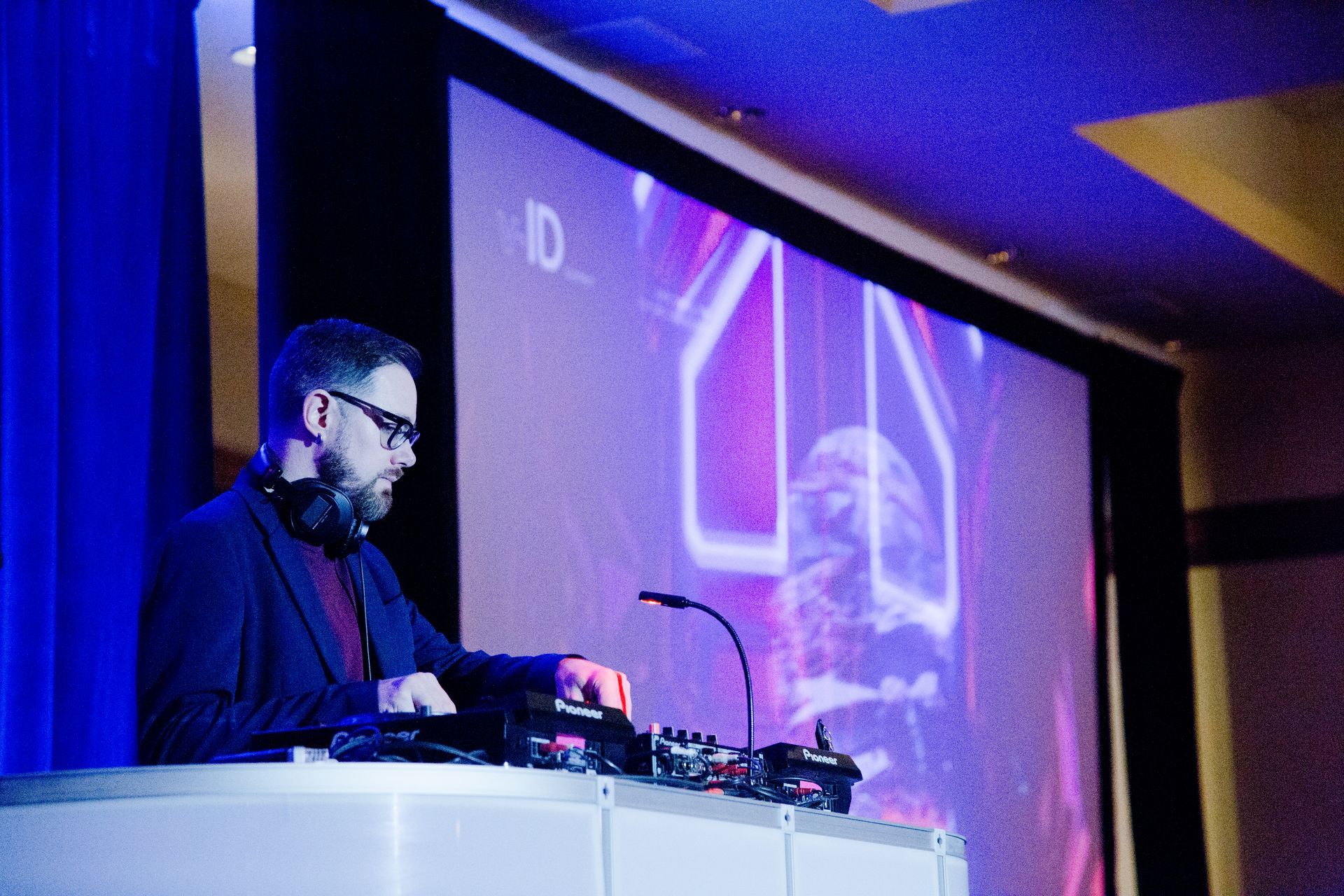 A person in a dark blazer with glasses operates a DJ controller on a white podium in front of a blue-lit stage.
