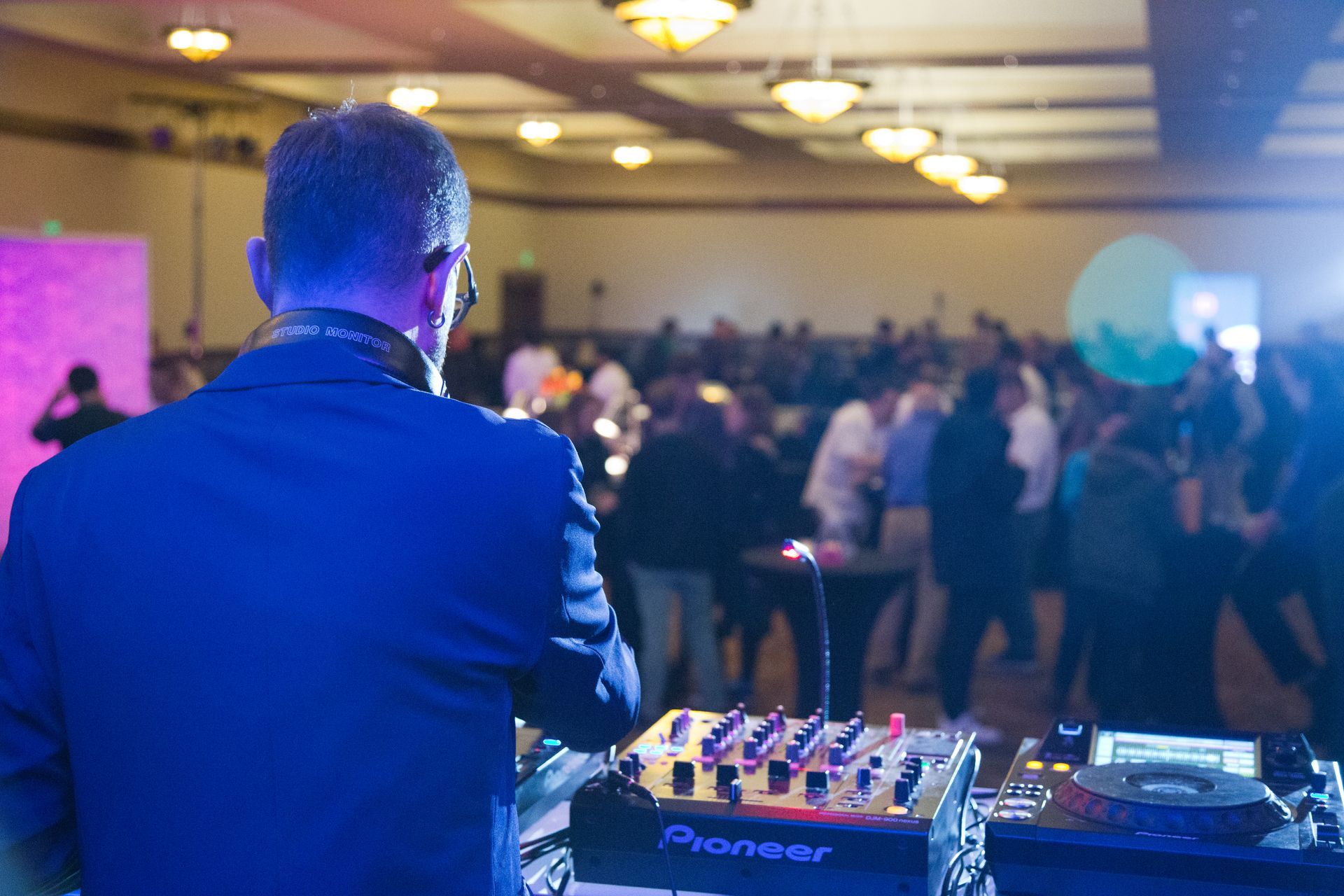 A DJ at a mixing console overlooking a dimly lit, crowded dance floor at an event venue.