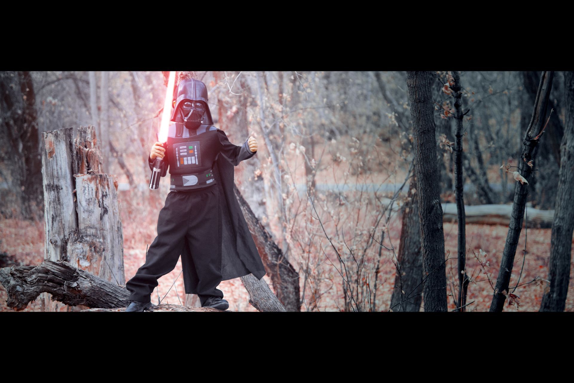 A child dressed in a Darth Vader costume holds a glowing red lightsaber in a wooded forest setting.