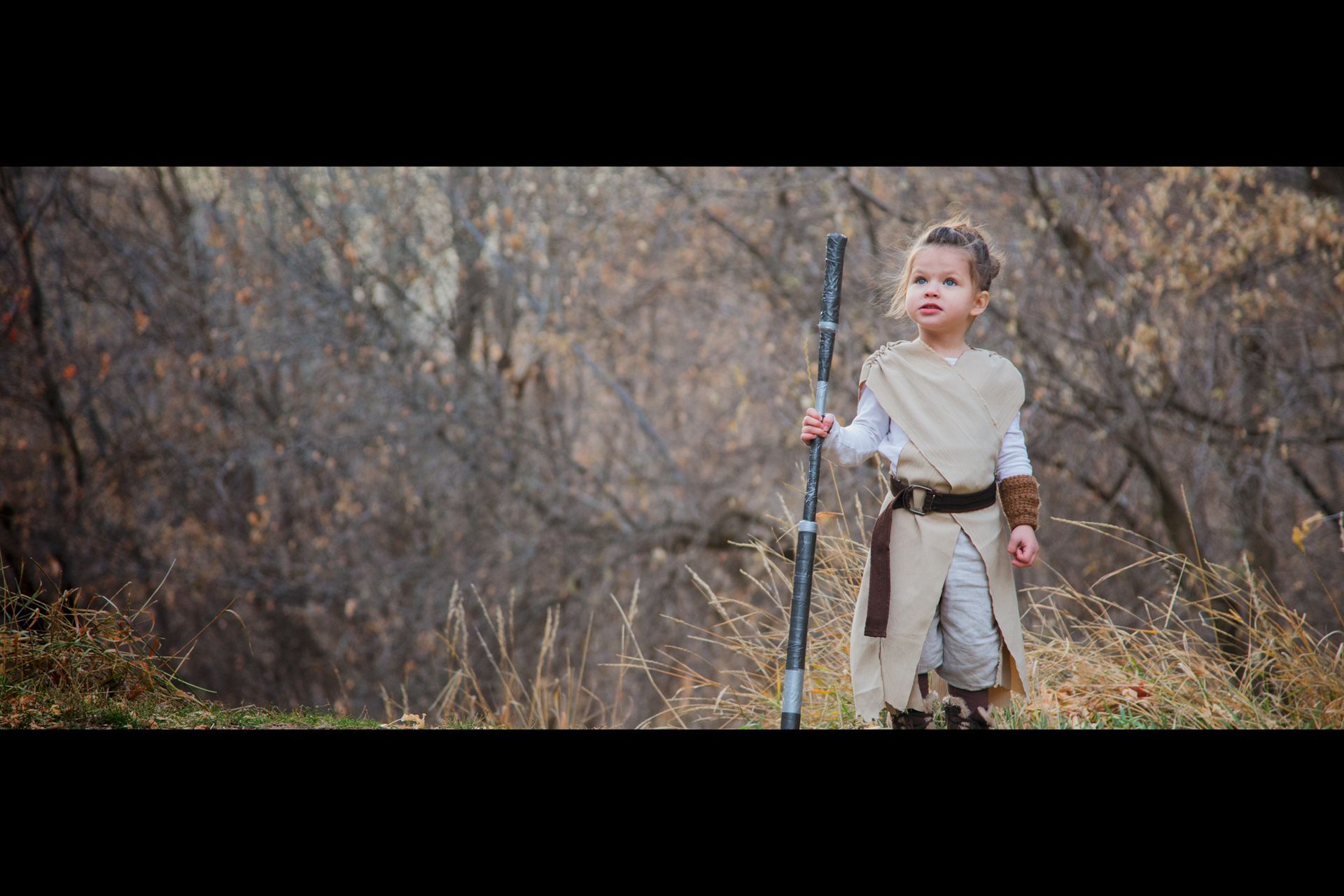 A child dressed as Rey from Star Wars stands in a wooded area, holding a staff.