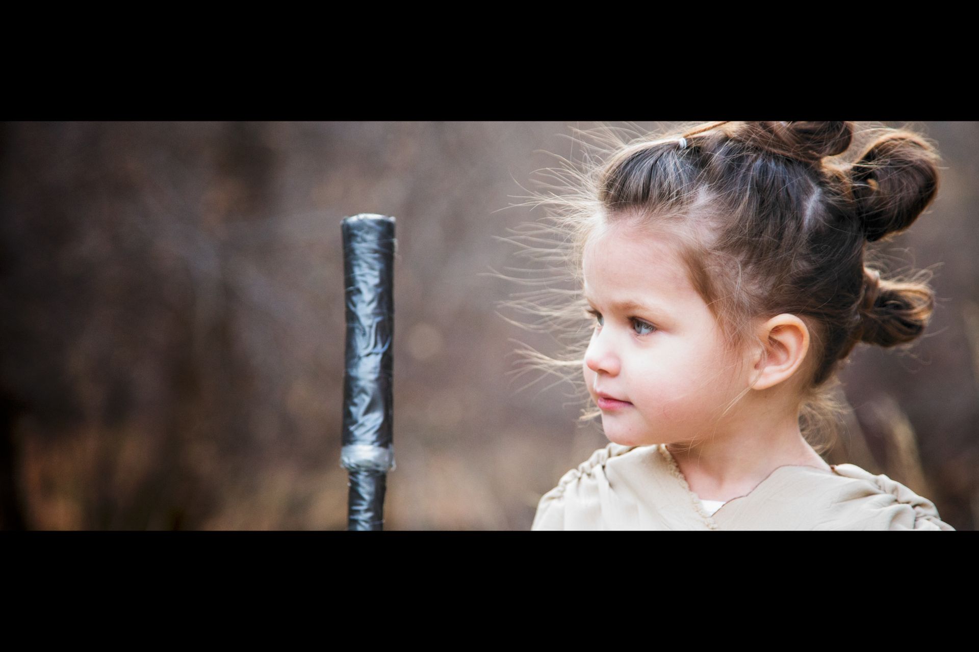 A child dressed as Rey from Star Wars, with iconic buns in their hair, looks intently at a prop staff.