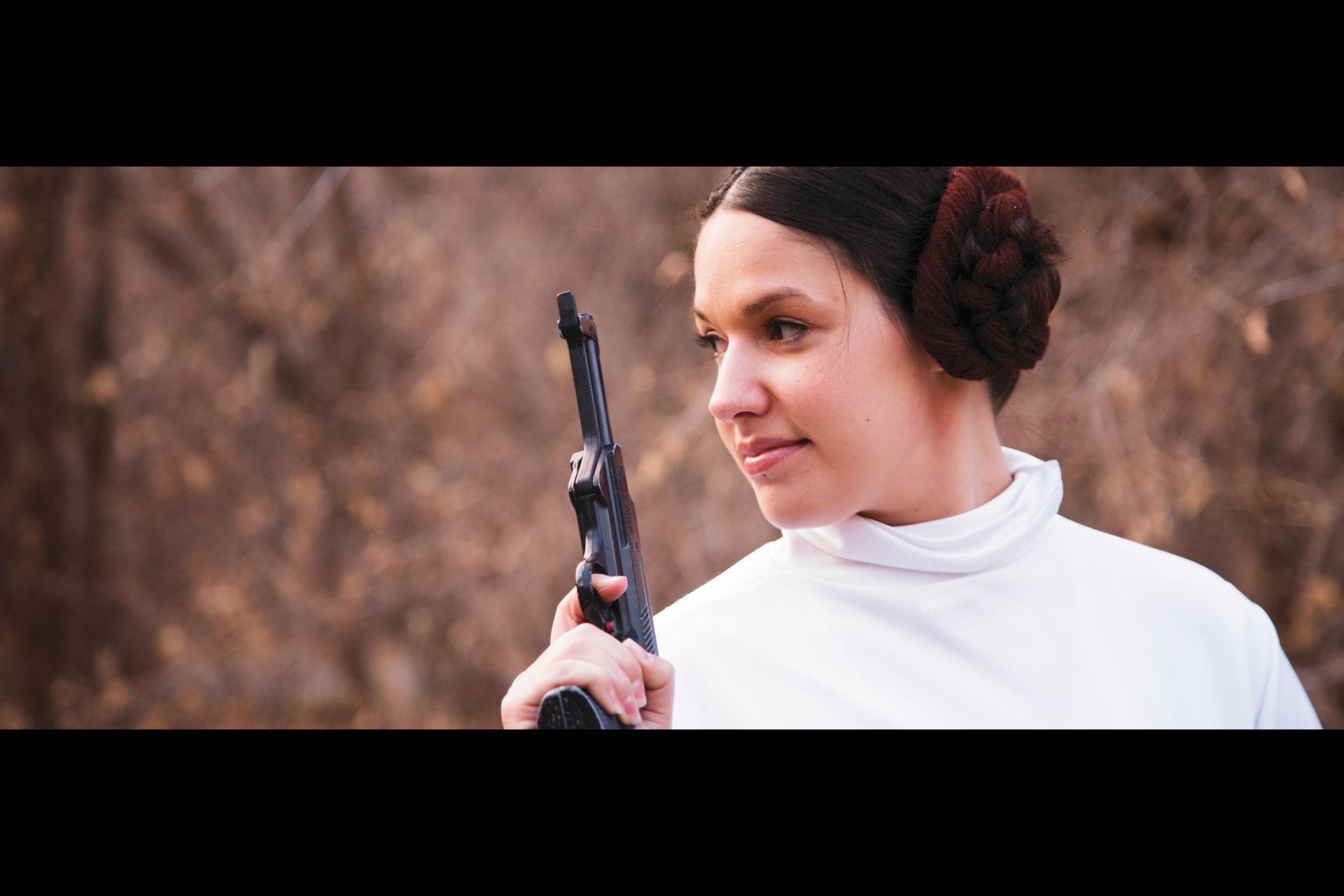 A person in a Princess Leia costume with iconic hair buns holds a prop blaster in an outdoor setting.
