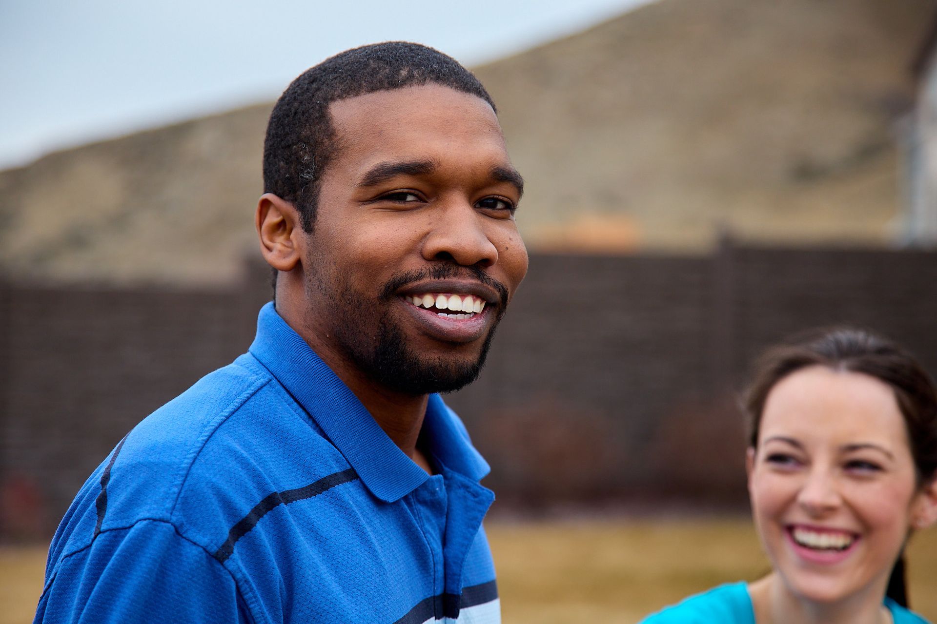 A man and a woman in blue tops smiling, with a blurred fence and hill in the background.