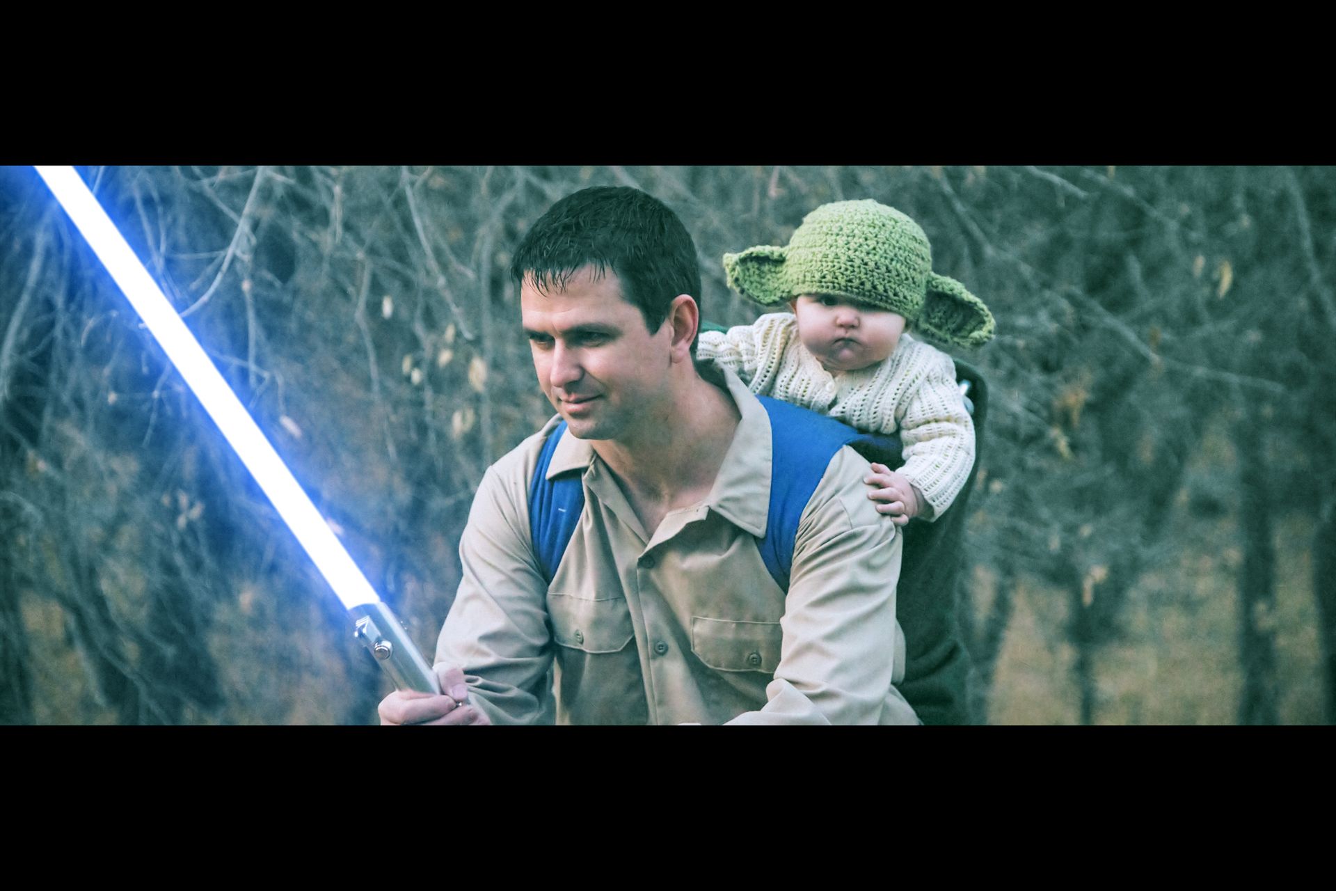A man carrying a baby in a Yoda-style knit hat on his back while holding a glowing blue lightsaber in a forest setting.
