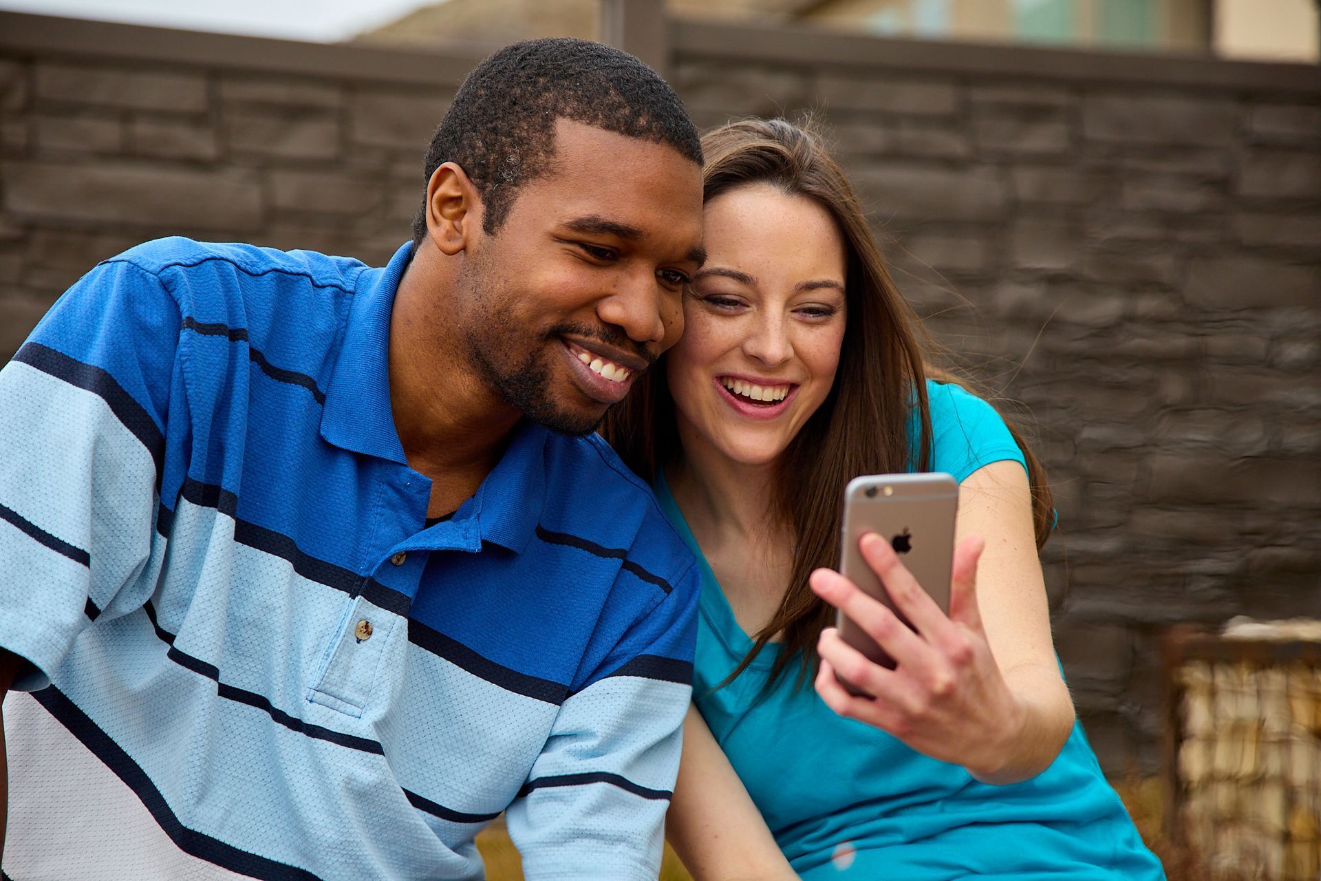 A smiling couple looks at a smartphone together against a dark, textured wall.