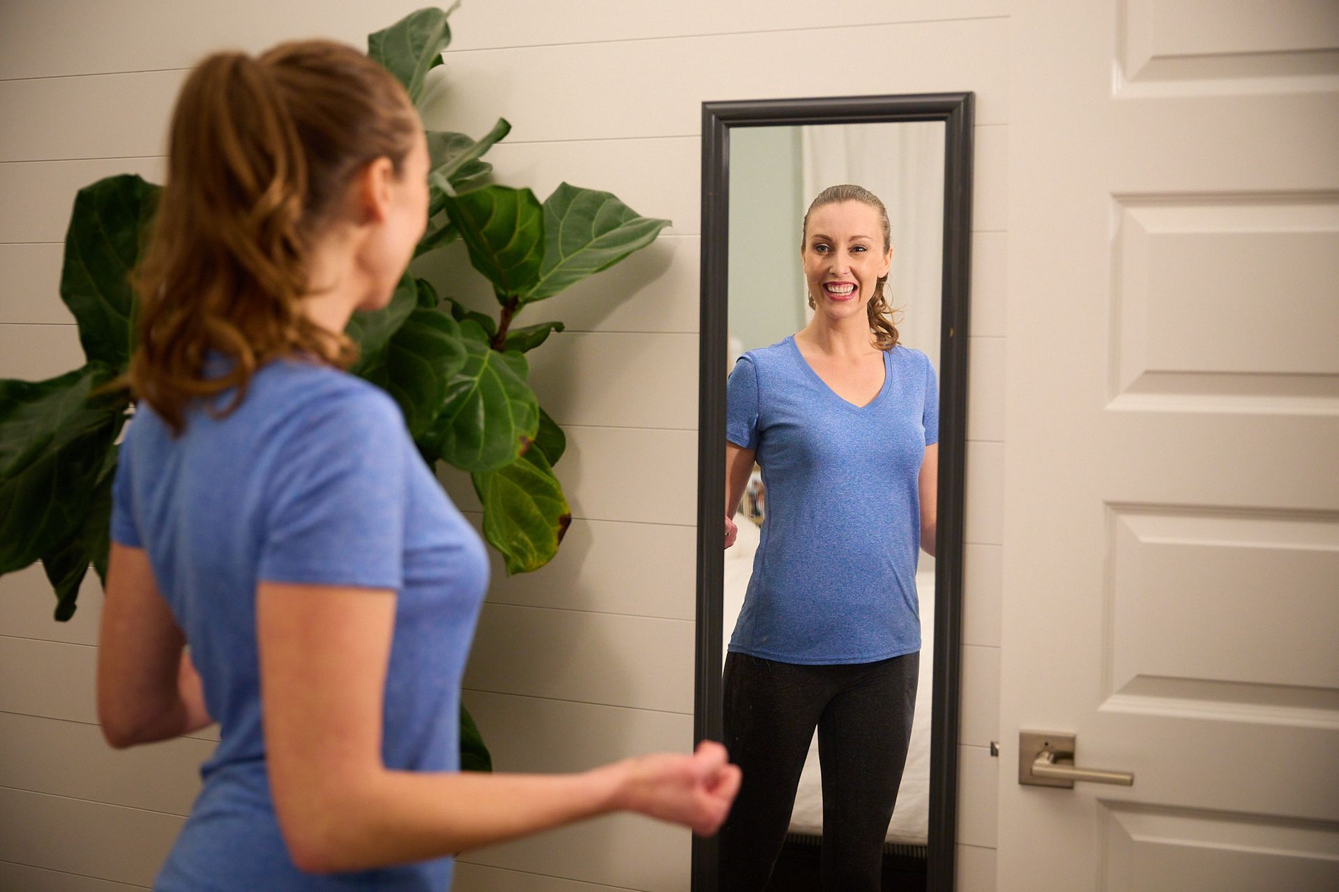 A person in a blue t-shirt and black pants smiles at their reflection in a full-length wall mirror near a large plant.
