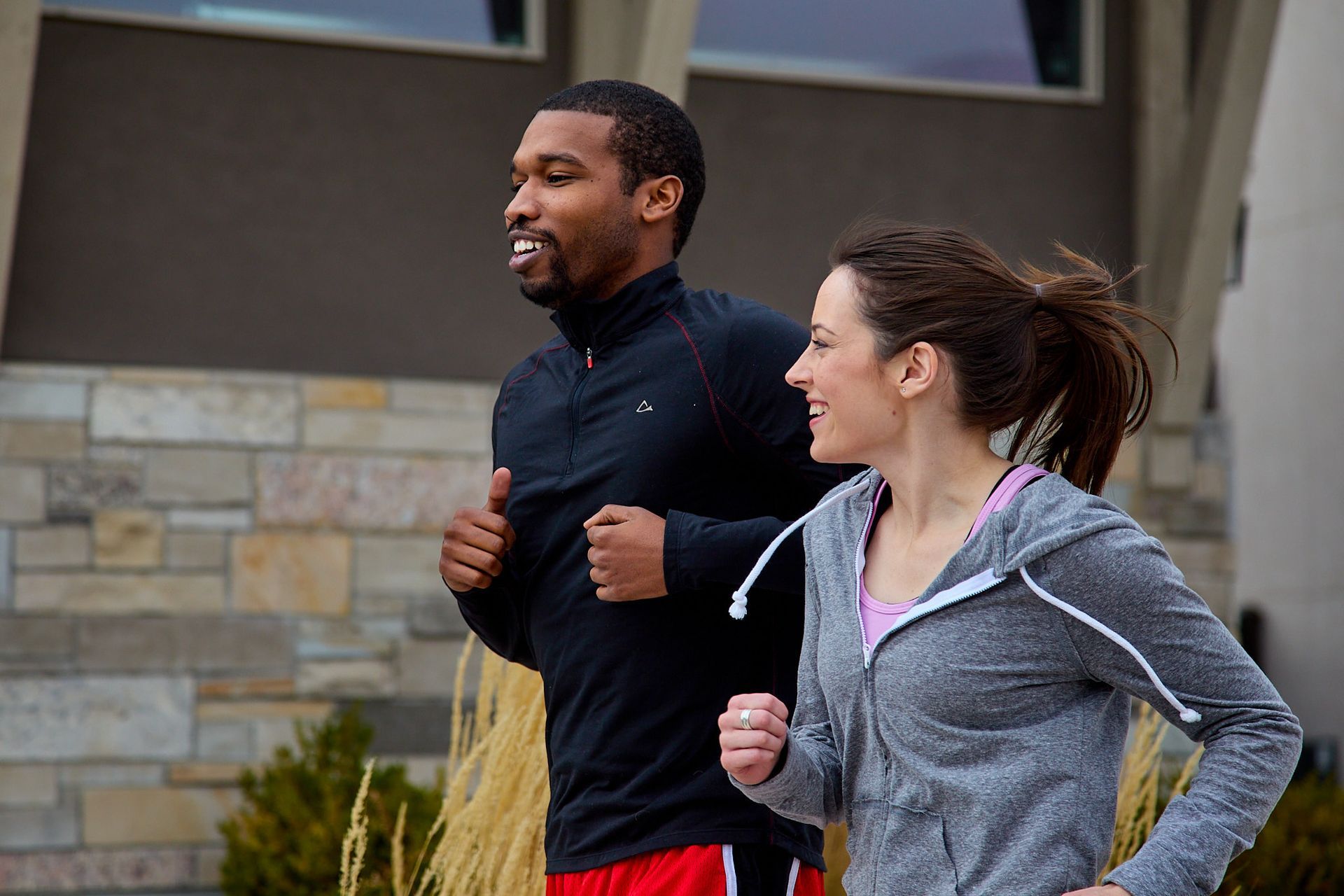 A person in a black long-sleeve shirt and another in a grey hoodie jogging together outdoors in front of a stone building.