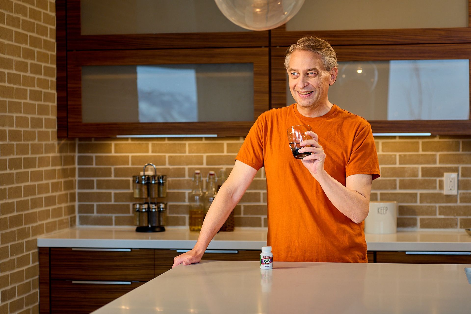 A person in an orange t-shirt holds a small glass in a modern kitchen with brick walls and wood cabinets.
