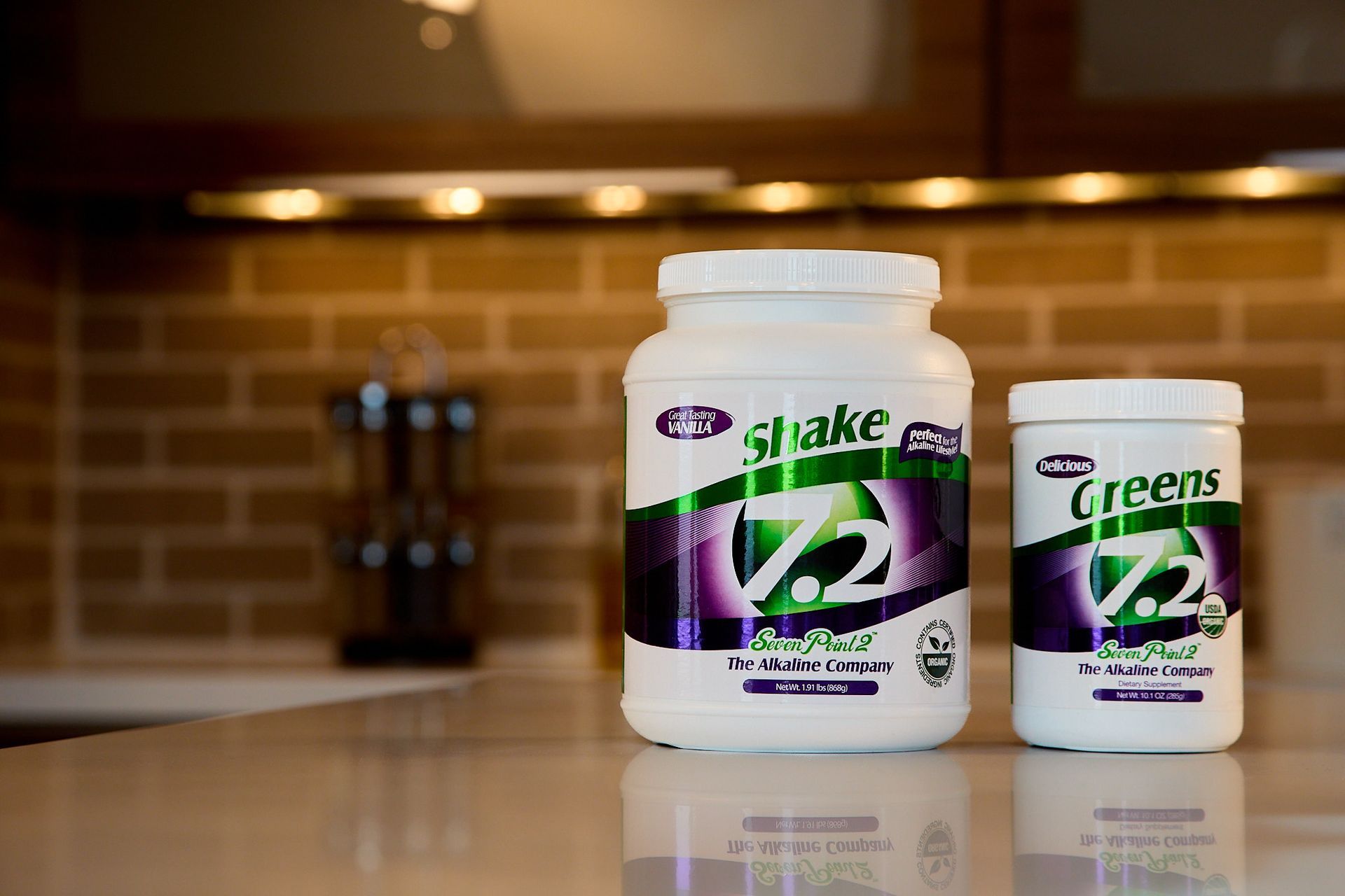 Two 7.2 brand supplement containers, one large Shake and one smaller Greens, sitting on a counter in a kitchen.