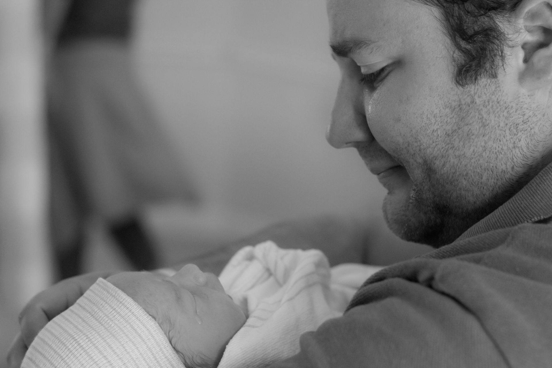 A man gazes tenderly at a newborn baby cradled in his arms.