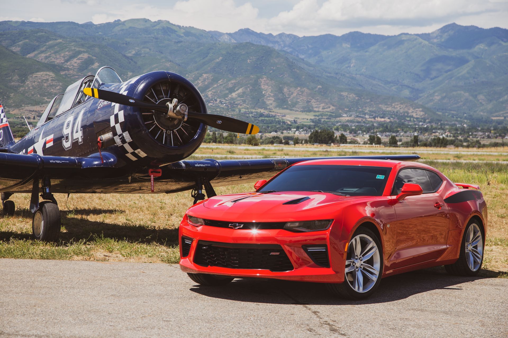 A red Chevrolet Camaro parked next to a blue vintage fighter plane on an airfield with mountains in the background.