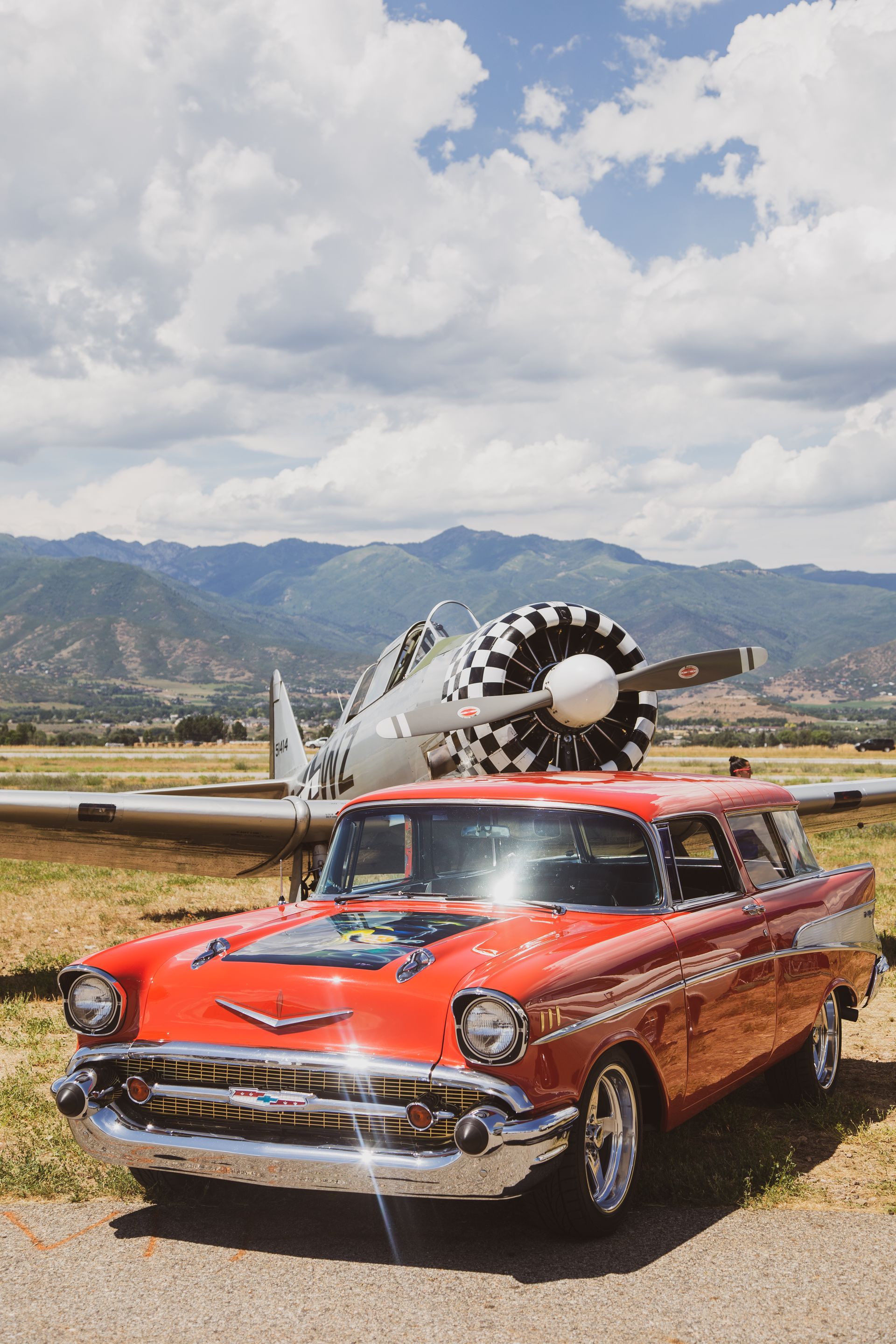 A bright red 1957 Chevrolet parked in a grassy field in front of a parked vintage airplane with mountains in the back.