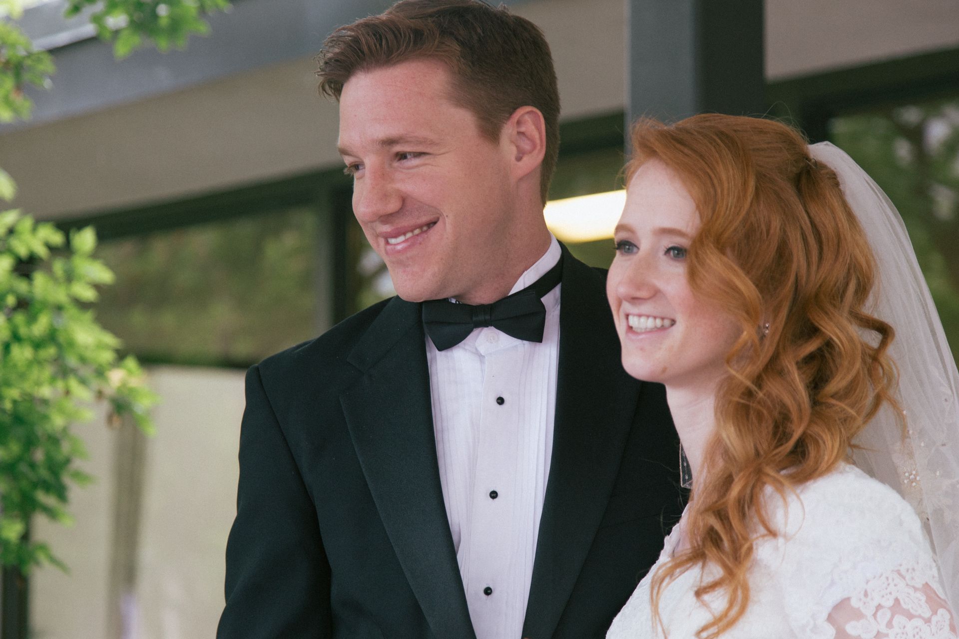 A smiling couple, dressed in a wedding tuxedo and a white bridal dress with a veil, posing together outdoors.