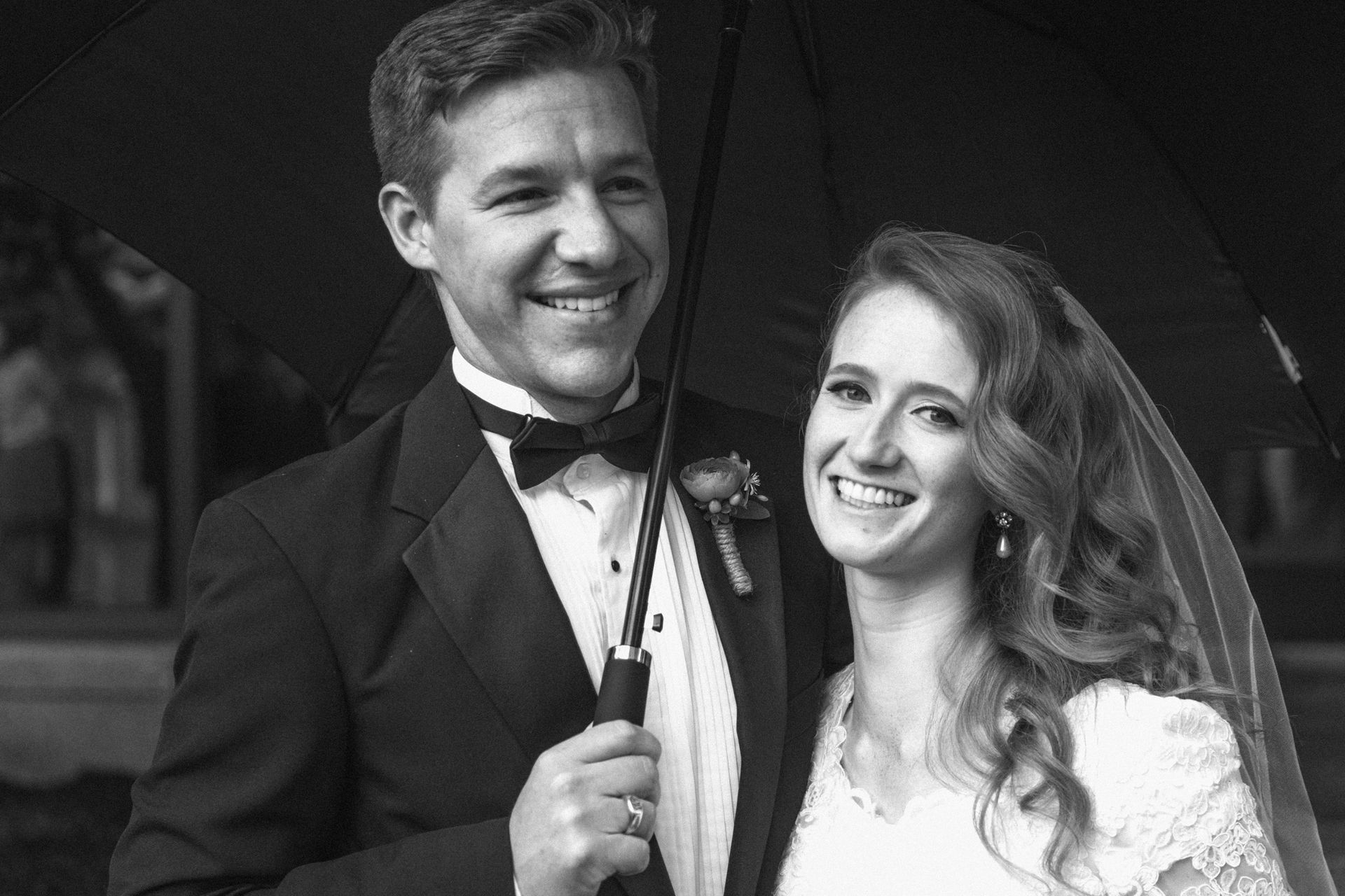 A smiling bride and groom in wedding attire stand together under a black umbrella in a black-and-white portrait.