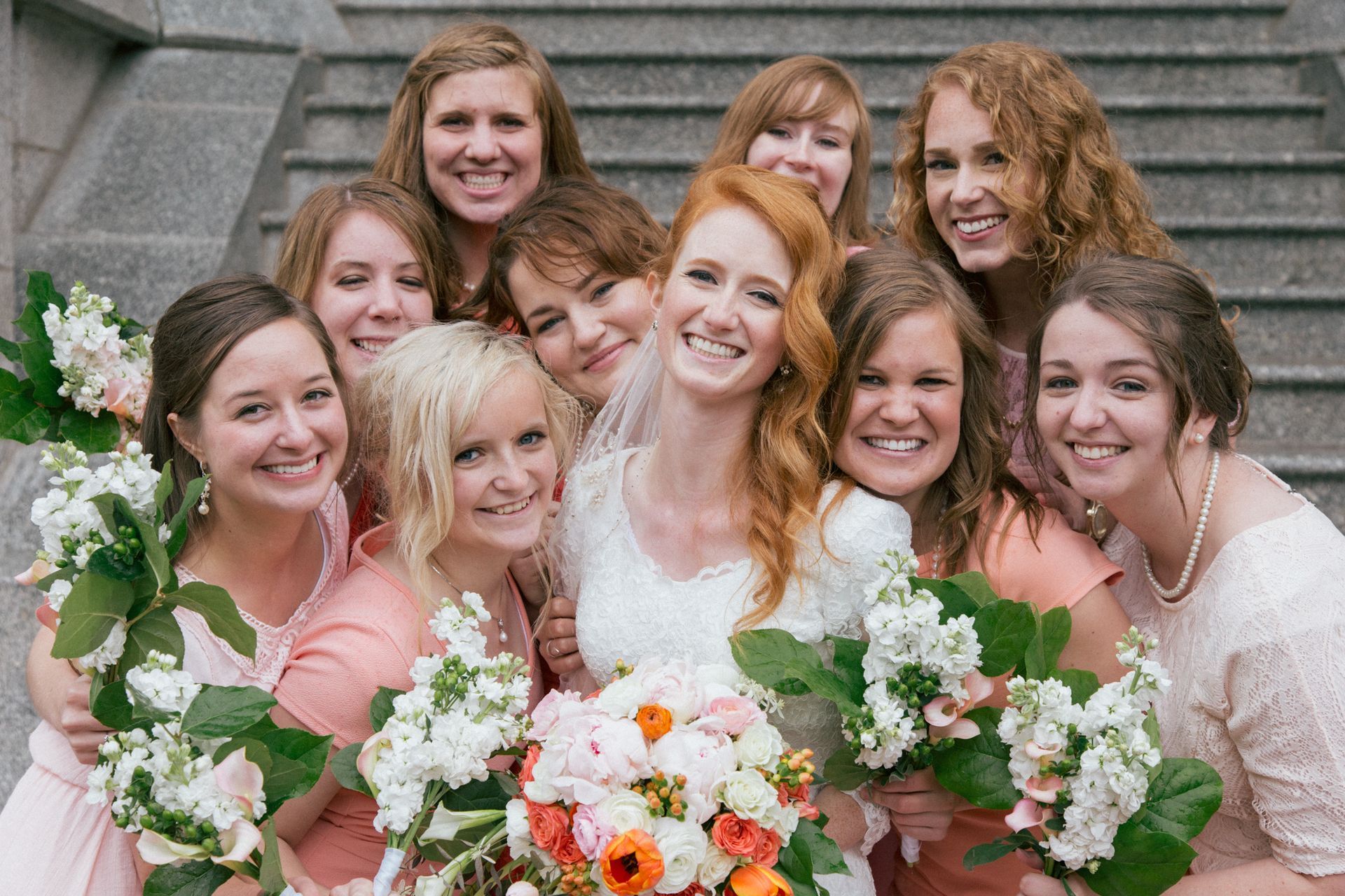 A bride and her bridesmaids in light-colored dresses pose together outdoors, smiling and holding bouquets of flowers.