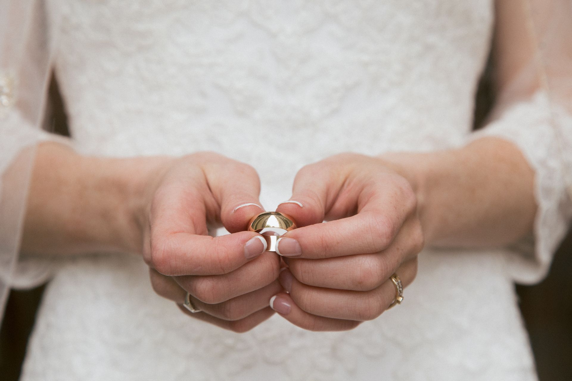 A close-up of a person in a white lace wedding dress holding a gold wedding band between their hands.