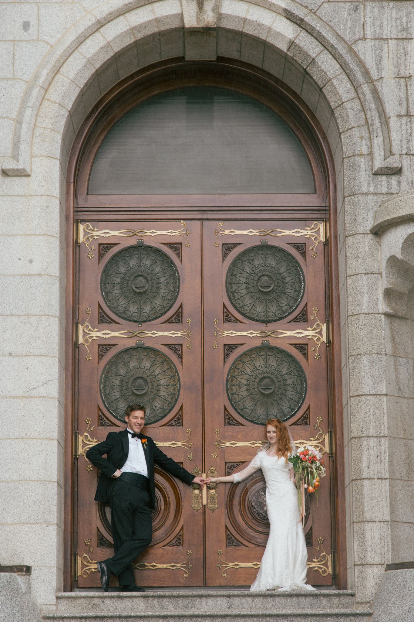 A bride and groom standing on stone steps in front of a large, ornate wooden double door, holding hands.