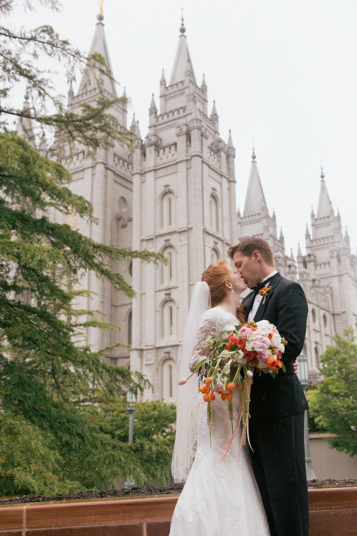 A couple kisses in front of the Salt Lake Temple, the bride holding a colorful bouquet and wearing a lace gown.