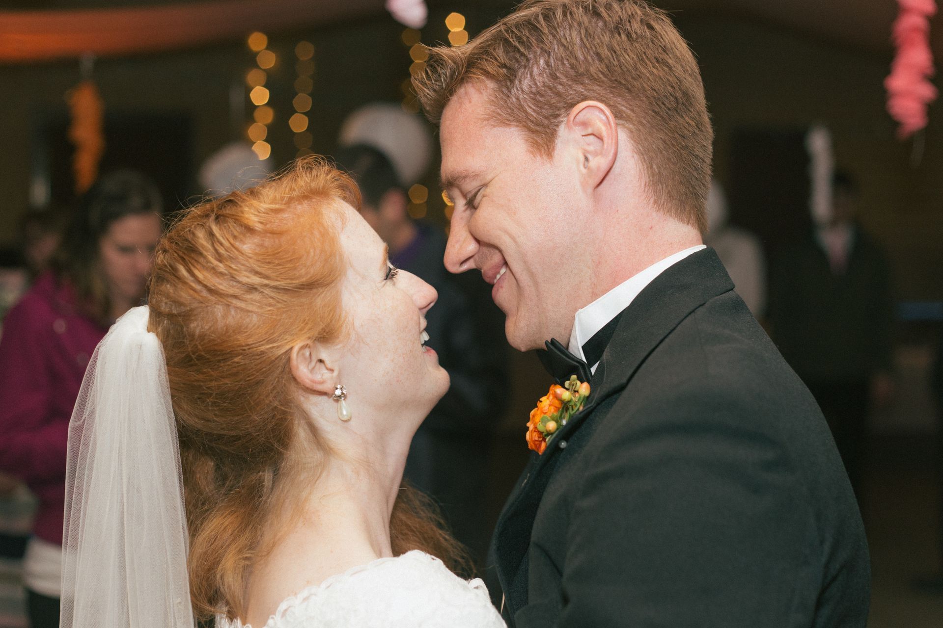A bride and groom in wedding attire smiling at each other during a dance in a dimly lit, decorated indoor setting.