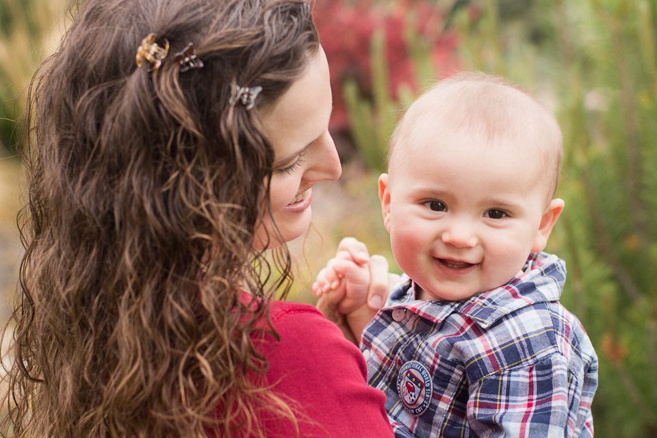 A person with long, curly brown hair and clips holds a smiling baby wearing a plaid shirt outdoors among greenery.