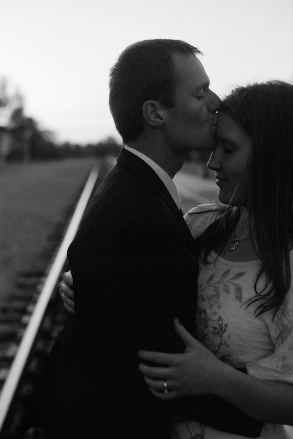 A man in a suit tenderly kisses a woman on the forehead while standing on a railroad track.