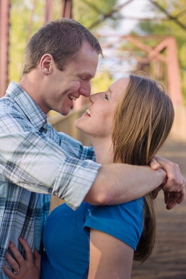 A man in a plaid shirt and a woman in a blue shirt embrace on a rustic bridge, looking toward each other and smiling.