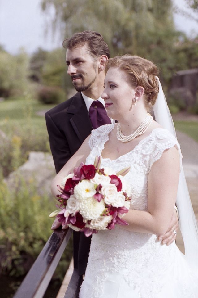 A couple in wedding attire poses together on a bridge, with the bride holding a bouquet of white and maroon flowers.