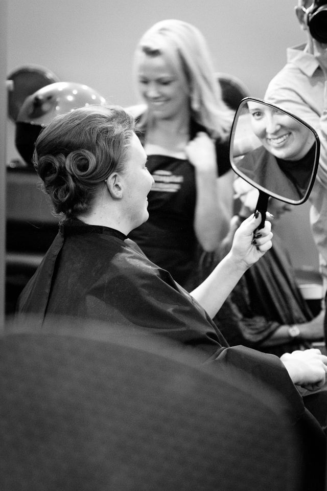 A person sitting in a salon chair looks into a handheld mirror, smiling at their newly styled hair while a stylist watches.