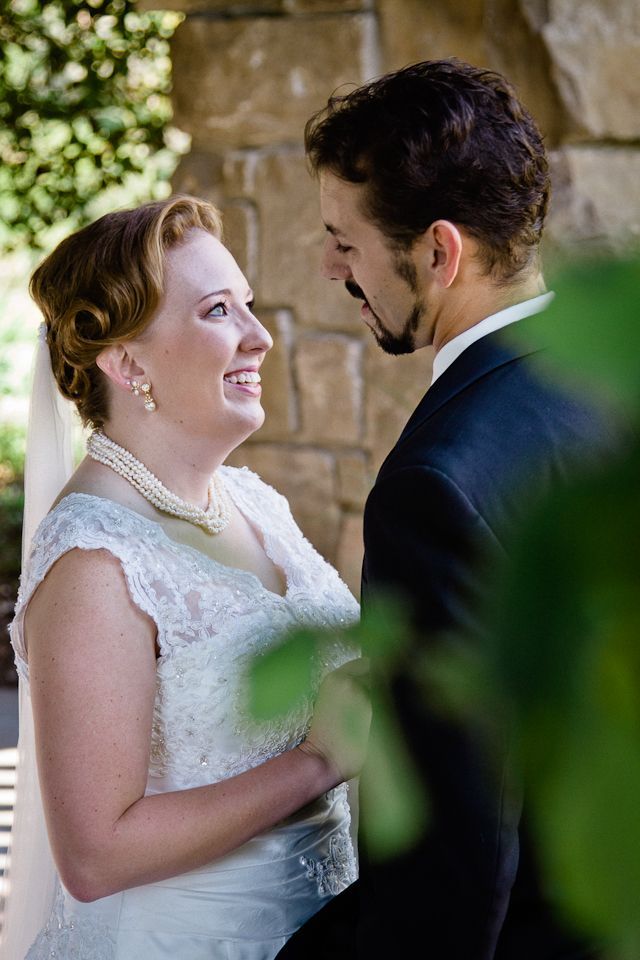 A bride in a lace dress and pearl necklace gazes lovingly at a groom in a dark suit before a rustic stone wall.
