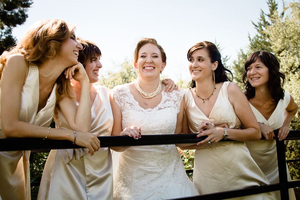 A bride and four bridesmaids in matching cream dresses laughing while leaning on a railing outdoors.