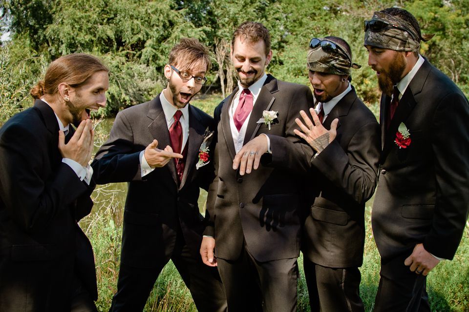 Four men in suits stand outdoors, excitedly gesturing toward a wedding ring on one man's hand.