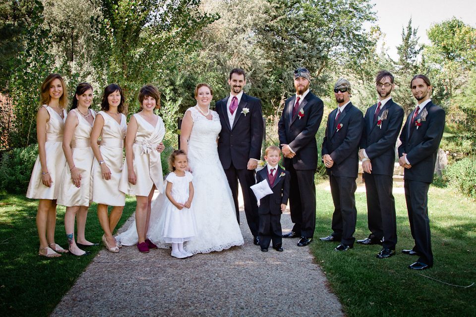 Wedding party posing for a group portrait in a park; bridesmaids in light dresses, groomsmen in suits, bride, and child.