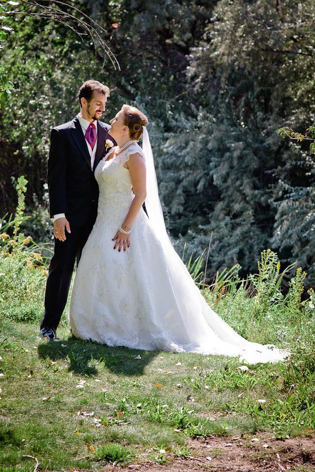 A bride in a white wedding gown and a groom in a dark suit stand facing each other in a grassy area with trees behind.