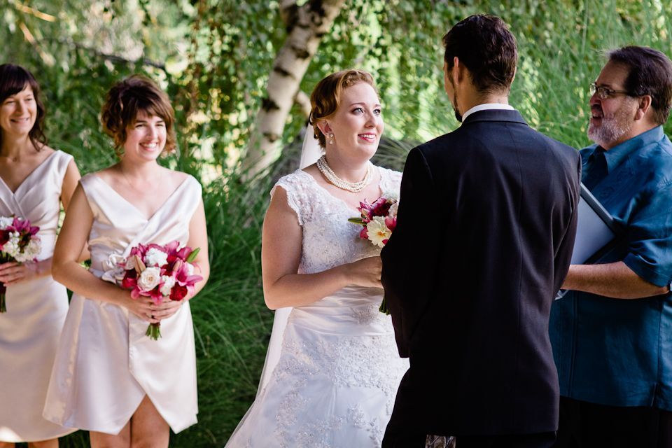 A bride and groom exchange vows outdoors, flanked by two bridesmaids in white dresses, with an officiant standing by.