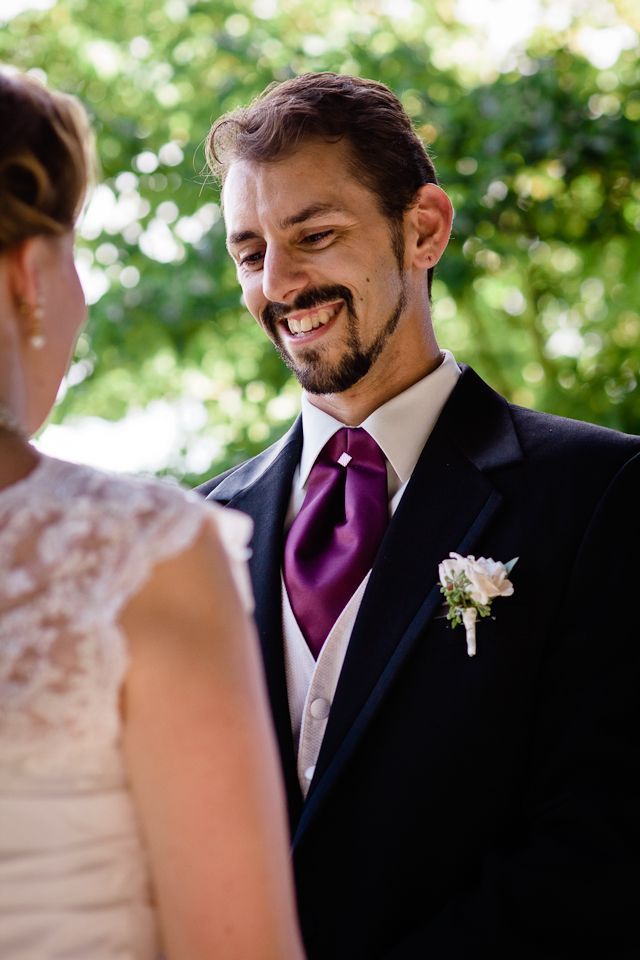 A groom in a black suit with a purple tie smiles lovingly at his bride during an outdoor wedding ceremony.